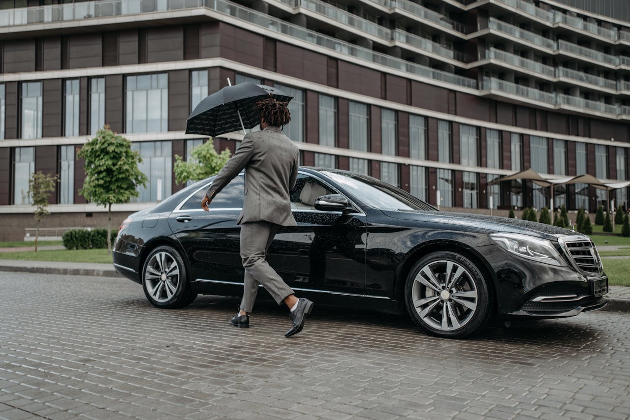 hero-img-01 Man holding umbrella opens car door on rainy day in front of modern building.
