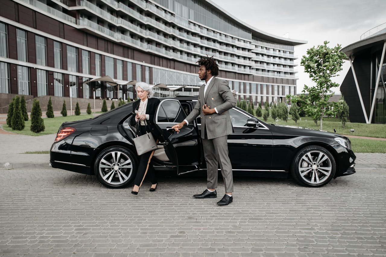 about-01 A professional chauffeur assists a businesswoman exiting a luxury car in front of a modern building.